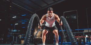Focused man in white tank top performing battle rope exercise in a modern gym.